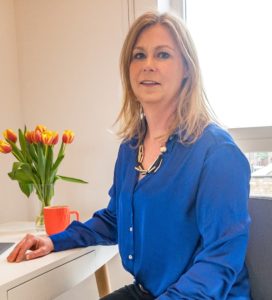 Photograph of Pinpoint Treasurer, Tina McEwan, sat at her desk. She is wearing a blue shirt and has a mug and vase of tulips in the background.